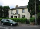 House with stained cream paint and dark slate roof.
Portico has pillars supporting chains above it.
Hedge, footway, and parked car in road.
Telephone pole.
Trees.