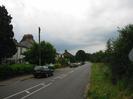 Road with trees and houses on left.
Parked cars on road.
Grass verge with trees and roadsigns on right.
Telephone pole.