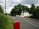 Road with grass verge on left.
Postbox and telephone pole in verge.
White house on far side of the road has low-pitch slate roof.
Traffic-calming `gate feature' visible in the distance.