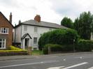 Road with footway and houses on the far side.
House on left is brick-built.
House on right has three storeys, and is white with dark slate roof.
Large hedges in front of white house partly obscuring a bus-stop sign.