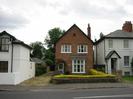 Row of houses on far side of road.
Centre house is built of bricks and has low wall and hedge around it's front garden.
Houses to left and right are white-painted with dark slate roofs.
Gravel driveway between houses.