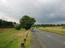 Road with grassy area on the left and field on the right.
Row of posts along the edge of the grass made from old telephone poles.
Trees and block of flats in the distance.