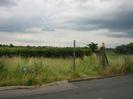 Road with grass verge and hedge on far side.
Entrance to footpath.
Chainlink fence on right surrounding allotment gardens.
Water-company markers in grass verge.
