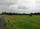 Road and footway on left.
Row of short posts beside footway.
Grassy slope drops towards the right, with playing fields and rugby posts in the distance.