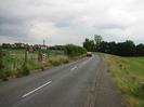 Road with allotment garden and chainlink fence on left.
GIVE WAY sign with three red bars on countdown board.
Road with footway on the right.
Grassy area far right, with row of low wooden posts made from old telephone poles.
Trees in the distance.