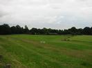 Slope down to area of rough grass.
Trees and club-huts in the distance.
Playing field on right.
Pile of rubbish left by encampment of travellers.