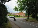 Looking north on Hitcham Road.
Dark bushes on left.
Cars and motorbike on road.
Houses beyond junction.
Roadsign warning of height limit.
Large tree in verge on right.