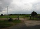 Entrance to sports club.
Concrete driveway with chainlink fence and metal gate.
Street light.
Low bushes.
Playing field in the background.