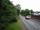 Looking east on Institute Road.
Grass verge and bushes on the left.
Car on road.
Pavement on far side, with gate leading to car showroom.
Low wall with fence on top on right, with cars behind.