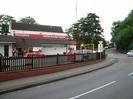 Looking west on Institute Road.
Station Garage Taplow on the left: white building with dark tile roof and red sign.
Cars on forecourt.
Wooden fence on low wall.
Junction with Station Road on right.
Approach Road in the distance, with trees and radio mast on the left and dark trees on the right.