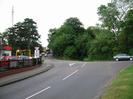 The junction of Institute Road, Station Road, and Approach Road.
Wooden fence with cars and buildings on left.
Yellow height-restricting gantry at entrance to car dealership.
Dark trees with radio mast.
Station buildings visible in the distance.
Trees on the far side of Station Road.
Grassy verge on right.