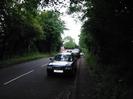 Looking south on Station Road.
Large trees on each side overhanging the road.
Parked cars.
Buildings at road junction visible in the distance.