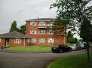 Hillmead Court flats.
Three-storey building with single-storey section on left.
Low-pitched roofs with gray tiles.
Lawn and flower-beds.
Parked cars.
Small tree in verge on right.