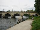 Maidenhead Bridge.
Small motor cruiser moored to near bank of River Thames.
Man reading on bench seat.
Large trees on right.