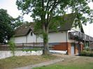 Maidenhead Rowing Club building.
Three-storey building with boathouse on ground floor.
Wooden balcony at first-floor level, with flagpoles.
Racing boats on racks.
Large trees.
Thames towpath in foreground.