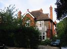 Cherry House.
Red brick house with white-painted windows and balcony.
Partly tile-hung on upper level.
Dark tile roof with decorative terracotta dragon on gable-end.
Tall chimneys.
Dark bushes along road.
Parked cars.