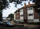 Bridge Court flats.
Three-storey building with reddish brick and white paint.
Gravel car-park.
Parked cars in road.
Trees overhanging road.