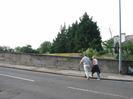 The eastern end of Maidenhead Bridge.
Roof of boat-shed visible over the parapet, with dark conifers beyond.
People crossing road.
Tall lamp post on right, with the roof of Skindles Hotel visible behind it.