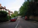 Ellington Road.
River Court flats behind bushes on the left.
Parked cars.
Fence and trees on the right.