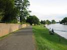 Looking south on River Road.
High sandy coloured brick wall on left with trees behind.
Grassy slope on right to River Thames.
People on riverbank.
Moored boats on river.