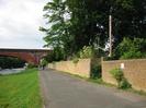 Looking north on River Road.
River on left with grassy bank to road.
Railway bridge in the distance.
High sandy-coloured brick wall on right with trees behind.
Telephone pole.