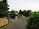 Looking south on River Road.
Sandy brick wall on left with trees behind.
Houses and people in the distance.
Bushes and grassy bank on right.