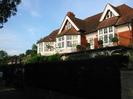 The Riverbank House seen across high hedge.
White-painted woodwork. Partly tile-hung at first floor level.
Red tile roof with small windows set into gable ends.
Iron railings around first-floor patio.