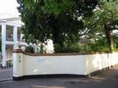 Folliotts.
White painted house with portico and covered veranda at first-floor level.
White-painted wall with brick capping.
White iron gates with globe lantern on gatepost.
Trees behind wall.