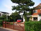 Pine Lodge.
House with white paint at ground-floor level and tile-hanging at first floor.
Awning over large picture window.
Gravel drive with brown wooden gates.
Scots Pine tree and cars in driveway.