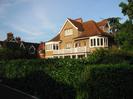 Sandy-coloured three-storey house with white painted windowframes and balcony.
Red tile roof.
High hedge between house and road.