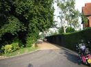 End of sealed road, with gravel drive beyond.
Large trees on left.
Clipped hedge on right with house visible over it.
Parked cars and motorcycle.