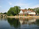 River Thames.
Eastern end of Maidenhead Bridge on left.
Large trees.
Maidenhead Rowing Club building: red bricks and green roller door at ground-floor level, white painted at first floor level, with wooden balcony. Two flagpoles on balcony. Glasshouse conservatory. Red tile roof.