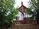 End wall of house seen through trees.
House has some white rendered areas and others done in red brick.
High wooden fence along road.