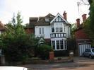 Two storey house with white paint and black details.
Dark tile roof with skylights and decorative ridge.
Hedge and trees in front garden.