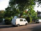 Road with parked car and van.
Long driveway on left leading to garage with white door.
Tree behind van, with houses beyond.