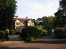 Large house of sandy brick with red bricks above windows and dark tile roof.
Gravel drive with hedges and trees.
Tarmac driveway on right leading to garage with white door.
Trees and telephone wires.