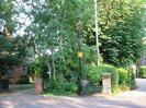 Road with Victorian-style street-light.
Yellow Neighbourhood Watch sign on street-light pole.
Gravel driveways with brick pillars.
Houses visible through trees.
Telephone pole.