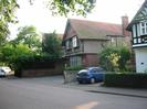 Road with houses on far side.
House in centre of picture has dark brick at ground floor level with bay window in black and white. Pebbledashed at first floor with box windows. Red tile roof with small window in gable end.
House on right is white with black timbers, white box window at first floor level.
Car in driveway. Concrete block wall. Low hedge.