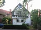 White house with black timber pattern on upper levels.
Bay window on ground-floor.
Two box windows on first floor.
Window under roof in gable end.
Tall chimney.
Low hedge alongside road.
Parts of other houses visible on each side.