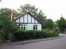 White bungalow with black timbers.
Bay window and smaller window on the right.
Low fence and hedge along road.
Trees behind.