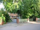 Road, pavement, and two driveways.
Drive on left has wooden gates and leads to two-storey brick house with bay window.
Drive on right has wooden gates on brick pillars and is laid with gravel.
Large trees between driveways.