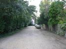 Ellington Gardens: gravel-surfaced road with trees on the left and wooden fence on the right.
Cars and house in the distance.