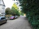 Gravel-surfaced road with house and parked cars on the left.
Trees overhanging on the right.