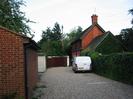 Gravelled parking area with brick garage on left.
Public footpath passes just to the right of the white pillar in the centre of the picture, but is not marked at this point.
Red tile-hung house behind parked van.
Ivy-covered wall on right.
Telephone pole.