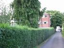 Privet hedge with tall conifer tree behind it.
Three-storey brick-built flats behind tree.
Tarmac driveway to Bath Road on right.