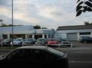 Road with VW car dealership on far side.
Cars on forecourt.
Low metal-clad showrooms and workshop buildings.