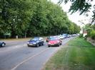 Road with queue of traffic.
Bank of large trees on the left.
Grass area on right with walls and gateposts.