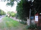 Wide grass verge with road on left.
Queue of cars on road.
Low walls along verge with shrubs behind walls.
House-name sign for Bridge House.
Public footpath sign pointing to the right.
Red brick pillar with decorative tile top and sign that says "MaST Consultancy & Training HERMITAGE HOUSE".
5 mph speed limit sign.