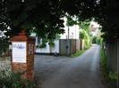 Tarmac driveway with overhanging trees.
Red brick pillar with signs "MaST Consultancy & Training HERMITAGE HOUSE" and 5mph speed limit.
White-painted house with wooden slat fence.
Hermitage House visible at end of driveway.
Public Footpath sign on extreme left points down driveway.