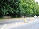 Road with pair of traffic islands, each with illuminated bollard.
Pavement and grass verge on far side of road.
Large trees.
Driveway between trees with brick gateposts.